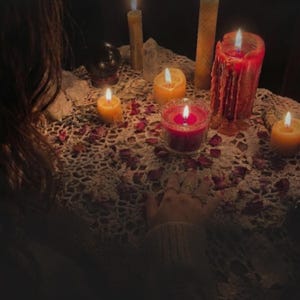 A person's hand is resting on a table covered with a lace doily, surrounded by lit candles and crystals, creating a warm and mystical atmosphere.