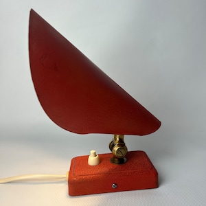 Può includere: Una lampada da tavolo in metallo rosso con un paralume triangolare e un giunto girevole in ottone. La lampada ha un interruttore bianco on/off e un cavo bianco.