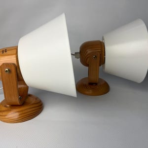 Może przedstawiać: Dwie drewniane lampy biurkowe z białymi stożkowymi kloszami. Lampy mają jasne drewniane wykończenie, regulowane klosze i okrągłą podstawę. Każda lampa jest podłączona do białego przewodu zasilającego.
