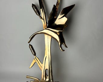 May include: A gold-toned metal wall sculpture depicting a stylized bird in flight, with reeds and cattails emerging from the water below. The sculpture has a textured, hammered finish.