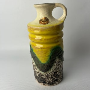 Puede incluir: Un jarrón de cerámica con asa, con una parte superior de color crema y un cuerpo amarillo con un diseño texturizado en verde oscuro y negro. El jarrón tiene una pequeña pegatina dorada cerca de la parte superior.