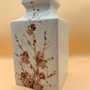 Può includere: Vaso in ceramica bianca di forma quadrata con un design floreale. Il vaso presenta un ramo delicato con fiori rosa e rossi.