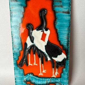 Puede incluir: Un azulejo de pared de cerámica con fondo azul y naranja y tres pájaros negros y blancos en relieve.