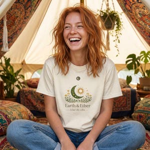 Op de afbeelding: Een crèmekleurig T-shirt met een halve maan, zon en bloemenontwerp met de tekst "Earth & Ether - Wear the vibes". De persoon draagt een blauwe spijkerbroek en zit in een tent.