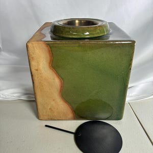 May include: A square ceramic oil burner featuring a glossy green crackle glaze on the top and front, transitioning into a natural tan unglazed side. It includes a circular metal insert at the top and a separate round black metal lid with a handle.
