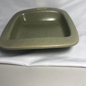 May include: A speckled, light green ceramic dish with a rectangular shape and rounded corners. The dish has a slightly raised rim and a smooth interior. The surface has small, dark speckles. A small label is visible on the top.