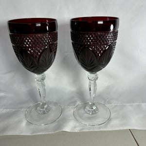 May include: Two wine glasses featuring deep red bowls with a textured diamond pattern on the upper section and a leaf-like motif on the lower section. Each glass has a clear, faceted stem and a round, transparent base, standing upright.