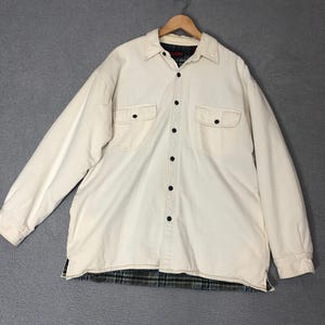 May include: Cream-colored, button-down shirt jacket with two chest pockets and black buttons. The jacket has a plaid lining visible at the hem and collar. The shirt is hanging on a wooden hanger.