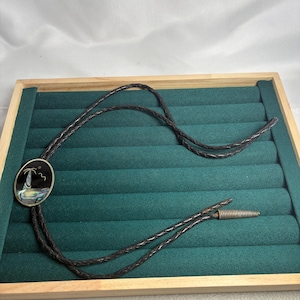 May include: A bolo tie featuring a dark oval pendant with an inlaid abalone shell design of a lighthouse and palm tree. The pendant is attached to a braided black leather cord that ends in silver-toned metal tips with a coiled spring detail.