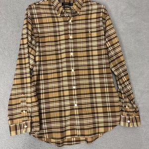 May include: A long-sleeved button-down shirt in a plaid pattern of brown, tan, and white. The shirt has a button-down collar, a small embroidered logo on the chest, and buttoned cuffs. The shirt is hanging on a wooden hanger.