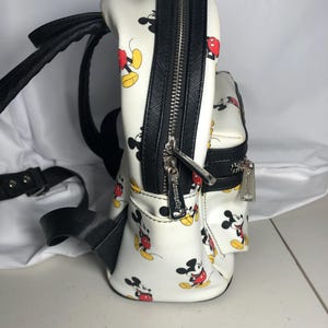 K&ouml;nnte beinhalten: Wei&szlig;er Mini-Rucksack mit einem sich wiederholenden Muster von Mickey Mouse in Rot, Gelb und Schwarz. Er hat schwarze Verzierungen, Riemen und Rei&szlig;verschl&uuml;sse mit silbernen Ziehern. Ein Accessoire zum Thema Disney.