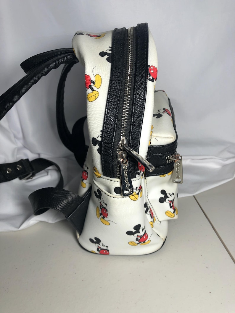 K&ouml;nnte beinhalten: Wei&szlig;er Mini-Rucksack mit einem sich wiederholenden Muster von Mickey Mouse in Rot, Gelb und Schwarz. Er hat schwarze Verzierungen, Riemen und Rei&szlig;verschl&uuml;sse mit silbernen Ziehern. Ein Accessoire zum Thema Disney.