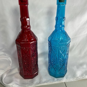 May include: Two tall glass bottles with cork stoppers, one in deep red and one in bright blue. Each bottle features a hexagonal body with an embossed leaf and geometric pattern, a narrow neck with a decorative ring, and a flared rim at the top.