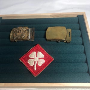 May include: Two rectangular brass belt buckles and one diamond-shaped red fabric patch with an embroidered white four-leaf clover. One buckle features an embossed eagle emblem, while the other is plain and smooth. The items are arranged together.