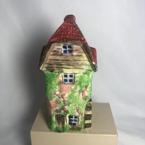 May include: A ceramic cottage figurine with a red roof and green walls covered in pink flowers. The cottage has small blue windows and a wooden door. The figurine is on a beige square base.