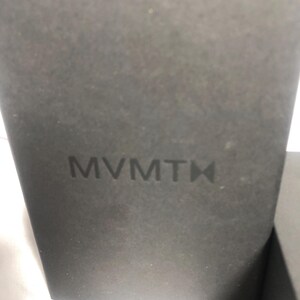 May include: A close-up of a dark grey box with the embossed brand name "MVMT" in the centre. The box has a textured surface and a minimalist design, suggesting product packaging.