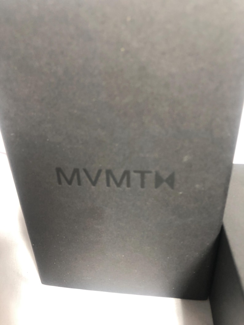 May include: A close-up of a dark grey box with the embossed brand name "MVMT" in the centre. The box has a textured surface and a minimalist design, suggesting product packaging.