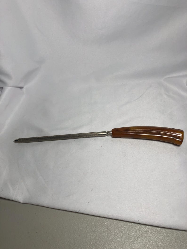 May include: A vintage knife sharpener. It features a long, slender steel rod and a curved handle. The handle is amber-coloured with a glossy finish. The sharpener is designed for sharpening knives and other cutting tools.
