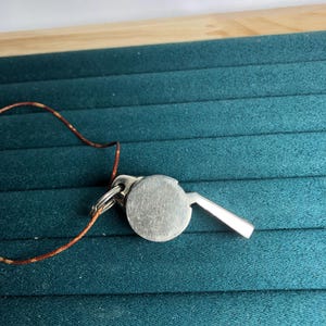 May include: A silver-colored whistle with a round body and a rectangular mouthpiece. It is attached to a small metal ring and a thin, brown cord. The whistle is set against a teal background.