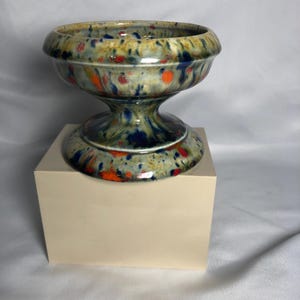 May include: A ceramic pedestal bowl featuring a glossy glaze with a speckled pattern in shades of blue, orange, and red over a cream-colored base. The bowl has a wide, rounded rim and a flared circular base, creating a distinct hourglass silhouette.
