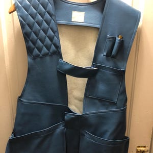 May include: A blue leather shooting vest with multiple pockets and a quilted shoulder pad. The vest is hanging on a wooden hanger. The vest has a classic design and appears to be made of high-quality materials.