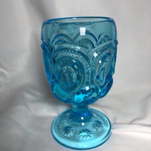 May include: A vintage, light blue glass goblet. The goblet features a detailed pattern of stars and crescent moons. The base and bowl are adorned with embossed floral designs. The stem is short and the glass has a glossy finish.