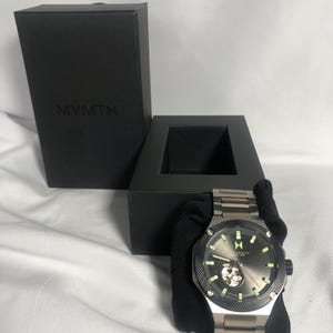 May include: A silver-toned MVMT watch with a dark grey face and a visible inner mechanism. The watch has a black bezel and luminous hands and markers. The watch is displayed with its black box, which has the MVMT logo.