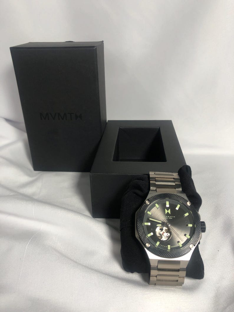 May include: A silver-toned MVMT watch with a dark grey face and a visible inner mechanism. The watch has a black bezel and luminous hands and markers. The watch is displayed with its black box, which has the MVMT logo.