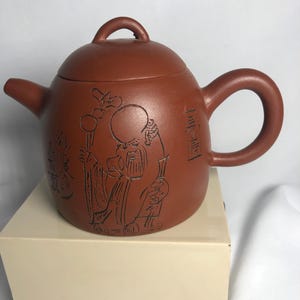May include: A reddish-brown ceramic teapot with a lid and handle. The teapot features an etched design of a figure holding a staff. The teapot is sitting on a beige box.
