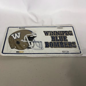 May include: A white license plate featuring a brown football helmet with a white "W" and the words "WINNIPEG BLUE BOMBERS" in blue and gold. The plate is rectangular with a blue border.