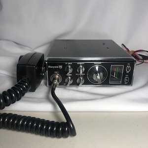 May include: A vintage Royce CB radio with a black microphone and coiled cord. The radio has a black face with silver knobs and a channel selector. The radio has a green and red signal meter and the word "Royce" is printed on the front.