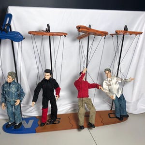 May include: Four male marionette dolls suspended from wooden control bars. The dolls wear various outfits including a denim jacket, a black and red jumpsuit, a red long-sleeve shirt with khaki pants, and a white button-down shirt with blue jeans.