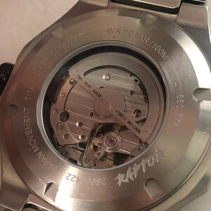 May include: Close-up of the back of a silver titanium watch. The watch has a transparent window showcasing the intricate inner workings. Text includes "MVMT", "JAPAN MOVEMENT TITANIUM", "WR 10 BAR/100M", and "RAPTOR".