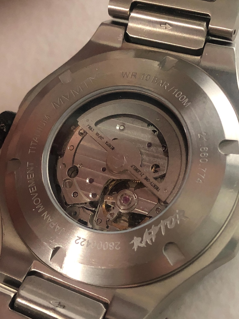 May include: Close-up of the back of a silver titanium watch. The watch has a transparent window showcasing the intricate inner workings. Text includes "MVMT", "JAPAN MOVEMENT TITANIUM", "WR 10 BAR/100M", and "RAPTOR".