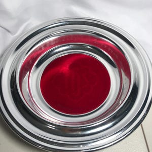 May include: A round, chrome-plated ashtray with a vibrant red velvet center. The ashtray features a multi-tiered design with a polished, reflective surface. The red center provides a striking contrast to the shiny chrome.