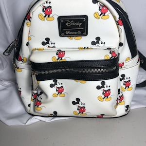 K&ouml;nnte beinhalten: Wei&szlig;er Disney Loungefly Mini-Rucksack mit einem sich wiederholenden Mickey Mouse-Muster. Der Rucksack hat schwarze Verzierungen, eine Vordertasche und einen schwarzen Rei&szlig;verschluss. Ein schwarzes rechteckiges Patch mit dem Disney Loungefly-Logo befindet sich vorne.