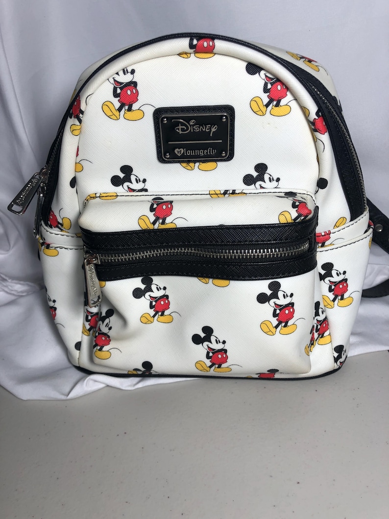 K&ouml;nnte beinhalten: Wei&szlig;er Disney Loungefly Mini-Rucksack mit einem sich wiederholenden Mickey Mouse-Muster. Der Rucksack hat schwarze Verzierungen, eine Vordertasche und einen schwarzen Rei&szlig;verschluss. Ein schwarzes rechteckiges Patch mit dem Disney Loungefly-Logo befindet sich vorne.
