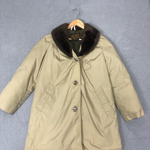 May include: A tan coat with a dark brown faux fur collar. The coat has a button-up front, two pockets, and long sleeves. It is hanging on a wooden hanger.