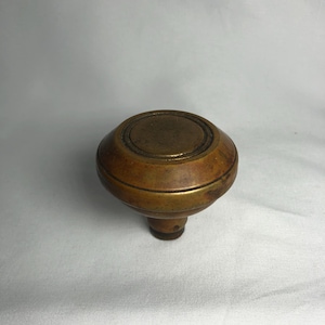May include: A vintage, round door knob with a brass finish. The knob has a slightly domed top with concentric circles and a cylindrical base. The brass has a warm, aged patina, suggesting it is an antique or vintage item.