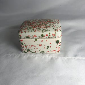May include: A rectangular ceramic box with a lid, featuring a white base color decorated with a speckled pattern of small red and green paint splatters across the entire exterior surface of both the lid and the base of the container.