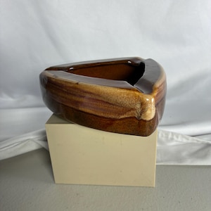 May include: A triangular ceramic ashtray featuring a glossy, multi-toned brown glaze with darker streaks and lighter beige highlights. The piece has a smooth, rounded rim and a deep, hollow center, showcasing a distinct gradient of earthy brown tones.