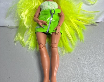 May include: A doll with long, bright neon yellow hair, brown skin, and large brown eyes. The doll wears a sleeveless white textured top and a lime green vinyl mini skirt featuring a silver zipper, a side buckle, and a metallic silver trim detail.