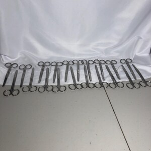 May include: A collection of silver-colored metal surgical scissors arranged in a horizontal row. Each pair of scissors features two circular finger loops and straight, pointed blades. The metal has a smooth, reflective finish throughout the set.