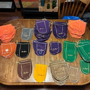May include: A collection of Crown Royal fabric drawstring bags in various colors including purple, orange, green, brown, yellow, gray, and tan. Each bag features the Crown Royal logo embroidered in gold thread, with some displaying additional graphics.