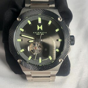 May include: A silver automatic watch with a black dial and visible movement. The watch has a textured bezel and a silver strap. The dial includes the text "AUTOMATIC 10BAR - 100M" and "JAPAN MOVEMENT".