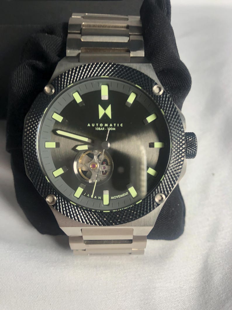 May include: A silver automatic watch with a black dial and visible movement. The watch has a textured bezel and a silver strap. The dial includes the text "AUTOMATIC 10BAR - 100M" and "JAPAN MOVEMENT".
