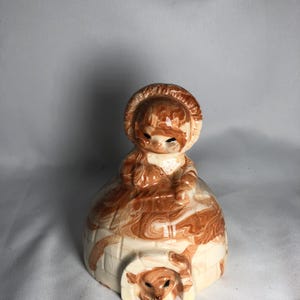 May include: A vintage ceramic figurine of a girl in a winter outfit, with a brown and white swirl pattern. The figurine has a small opening at the base with a small animal inside. The figurine is a decorative item.