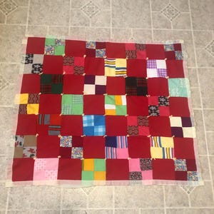 May include: A square patchwork quilt featuring a grid of solid red fabric squares alternating with smaller multi-colored patterned fabric blocks. The quilt is constructed with visible white yarn ties at the corners of the squares and has a light-colored border.