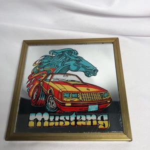 May include: A framed mirror featuring a vintage Mustang car graphic. The car is red and yellow with blue accents, and a blue horse head above it. The word "Mustang" is displayed in colorful block letters.