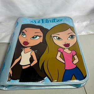May include: A light blue zippered binder featuring the Lil Bratz logo at the top. The front cover displays a graphic illustration of two cartoon characters with long hair, one with dark brown hair and one with blonde hair, wearing casual clothing.